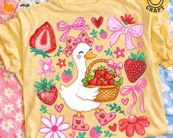 May include: Yellow t-shirt with a whimsical strawberry and goose design. The graphic features a white goose carrying a basket of strawberries, surrounded by bows, hearts, and flowers. The text "tiensoncraft" and a smiley face logo are visible.