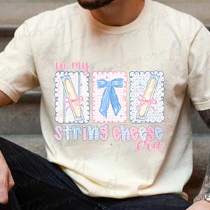 May include: Cream-colored t-shirt with a graphic design featuring the text "in my string cheese cra." The design includes three rectangular frames with string cheese and bows in pink and blue.