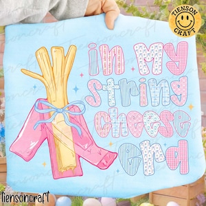 May include: Pale blue t-shirt featuring the text "In My String Cheese Era". The design includes a cartoon string cheese stick wearing a pink jacket and a blue bow. A fun, whimsical design, perfect for casual wear.