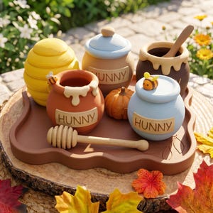 Può includere: Un set di quattro barattoli per miele in ceramica su un vassoio marrone, che include un barattolo giallo a forma di alveare, uno beige con coperchio blu, uno marrone scuro con mestolo in legno e due barattoli arancioni e blu etichettati HUNNY.