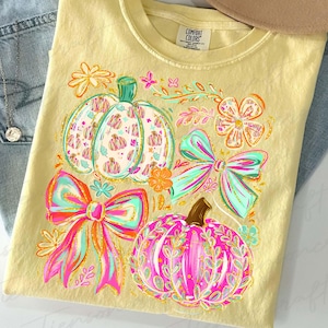May include: A yellow Comfort Colors t-shirt featuring a colorful fall design. The design includes pumpkins, bows, and floral elements in shades of pink, teal, orange, and gold. The shirt is laid flat on a surface.