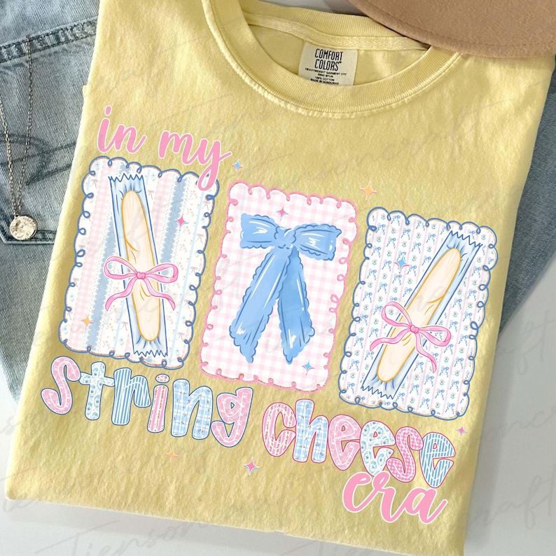 May include: Yellow Comfort Colors t-shirt with a graphic design featuring the text "in my string cheese era." The design includes illustrations of string cheese and a blue bow. The shirt is laid on top of blue jeans.