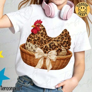 May include: White t-shirt featuring a hen with a leopard print design, sitting in a woven basket with a cream-colored bow. The hen has a red comb and yellow beak. The shirt has the text "tiensoncraft" on it.