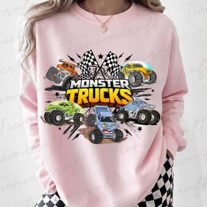 May include: Pale pink sweatshirt with a vibrant graphic of five monster trucks. The design includes a shark, dinosaur, and other truck styles, with the words "MONSTER TRUCKS" and checkered flags. The sweatshirt is a casual wear item.