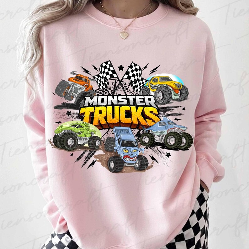 May include: Pale pink sweatshirt with a vibrant graphic of five monster trucks. The design includes a shark, dinosaur, and other truck styles, with the words "MONSTER TRUCKS" and checkered flags. The sweatshirt is a casual wear item.