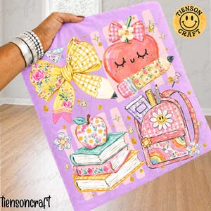 May include: A light purple blanket featuring a school-themed graphic print with a large yellow plaid bow, a smiling apple with a pencil, a stack of three books, and a pink backpack with a daisy, all accented by small gold stars and dots.