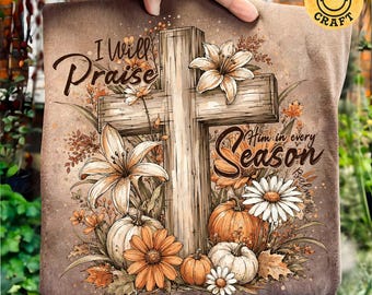 May include: A brown fabric item featuring a graphic of a wooden cross surrounded by orange and white lilies, daisies, and pumpkins. The text reads I Will Praise Him in every Season and Psalm 146:2 in a dark brown script font across the design.