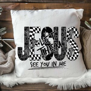 May include: White square cushion with the words "JESUS LET THEM SEE YOU IN ME" in black, with checkered and cross designs. The cushion is on a wooden surface.