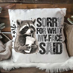 May include: White throw pillow featuring a raccoon drinking from a cup with a straw. The pillow has the text "SORRY FOR WHAT MY FACE SAID" in a mix of fonts and patterns, including checkered and leopard print.