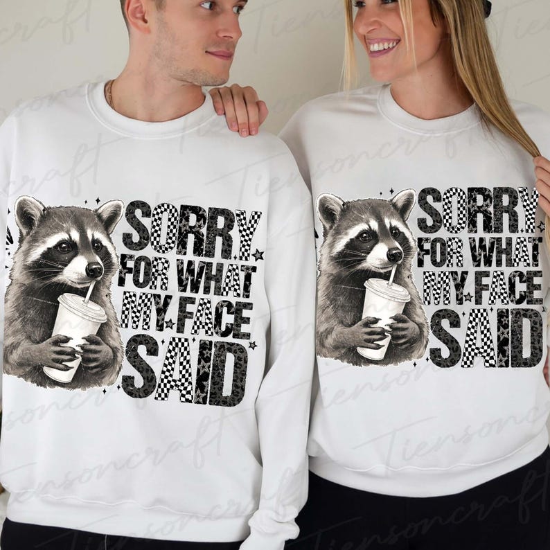 May include: White sweatshirts with a raccoon graphic holding a drink. The text "SORRY FOR WHAT MY FACE SAID" is printed below the raccoon in a mix of fonts and patterns, including checkerboard and stars.