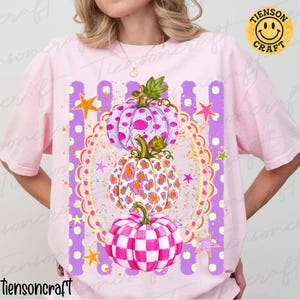 May include: A light pink t-shirt featuring a graphic of three stacked pumpkins inside a decorative yellow scalloped oval frame. The top pumpkin is light purple with spots, the middle has an orange leopard print, and the bottom is pink checkered.