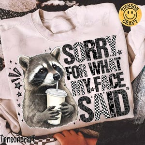 May include: A light pink sweatshirt featuring a raccoon drinking from a cup with a straw. The text "SORRY FOR WHAT MY FACE SAID" is printed in a mix of checkered and leopard print patterns. The sweatshirt has a crew neck.