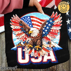 May include: Black t-shirt with a patriotic design. It features a bald eagle with wings decorated in red, white, and blue patterns, set against an American flag and a mountain backdrop. The word "USA" is prominently displayed in red and blue.