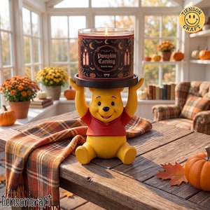 May include: A yellow bear figurine with a red shirt holding a black candle holder base. A cylindrical candle with a dark label featuring the text Pumpkin Carving, bats, and moons sits on the base. The bear has a smiling face and raised arms.