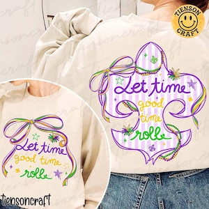 May include: Cream-colored sweatshirt featuring a purple, green, and yellow fleur-de-lis design with the text "Let time good time rolle." The design includes a bow, stars, and a string of beads. A yellow smiley face logo is in the upper right corner.