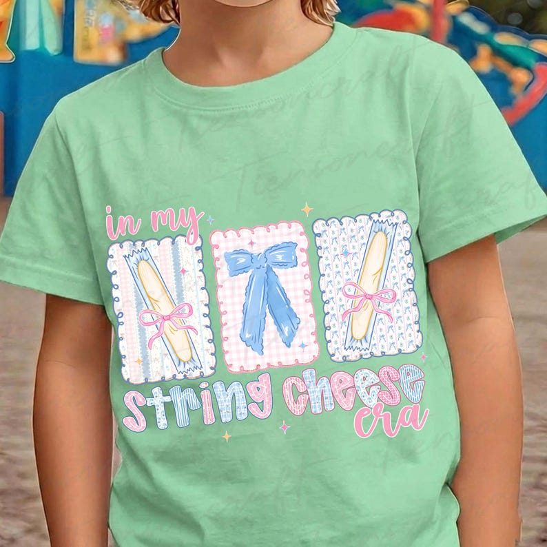 May include: A light green t-shirt featuring the phrase "in my String Cheese era" in a playful font. The design includes three rectangular illustrations of string cheese and a blue bow. The shirt is made of a soft material.