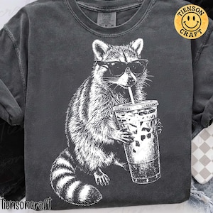 May include: Dark grey sweatshirt featuring a white graphic of a raccoon wearing sunglasses and drinking from a clear iced coffee cup. The text "tiensoncraft" is at the bottom, and a yellow smiley face logo is on the upper right.