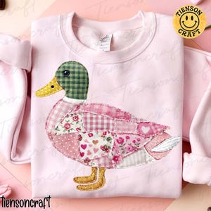 Könnte beinhalten: Hellrosa Sweatshirt mit einem Patchwork-Enten-Design. Die Ente hat einen grün karierten Kopf, einen gelben Schnabel und einen Körper aus rosa Blumen- und Karostoffen. Auf dem Sweatshirt steht unten der Text "Tiensoncraft".