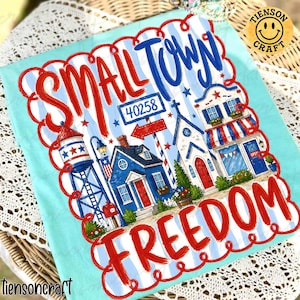 Könnte beinhalten: Hellblaues T-Shirt mit patriotischem Design. Es zeigt die Worte "SMALL TOWN FREEDOM" in roter und blauer Schrift. Das Design umfasst Illustrationen eines Wasserturms, Häusern und einer Kirche, mit der Postleitzahl 40258.
