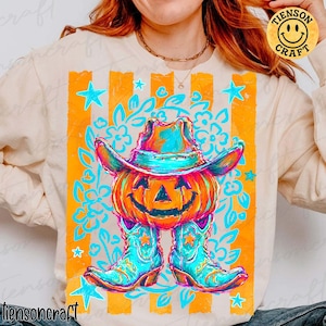 May include: A cream-colored long-sleeve sweatshirt featuring a graphic print of an orange pumpkin wearing a turquoise cowboy hat and matching turquoise cowboy boots. The design includes orange vertical stripes, blue stars, and floral patterns.