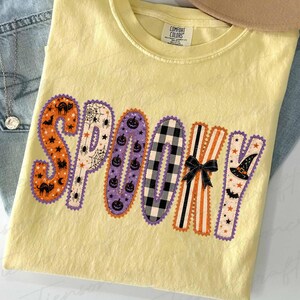 Może przedstawiać: Jasnoż&oacute;łty t-shirt z kr&oacute;tkim rękawem z napisem SPOOKY wydrukowanym dużymi, ozdobnymi literami. Każda litera ma inny wz&oacute;r, w tym pomarańczowy z czarnymi kotami, pajęczyny, fioletowe dynie, czarno-białą kratę oraz paski.