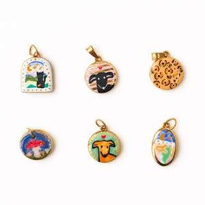 May include: Six gold-toned metal pendants featuring colorful enamel designs: an arched cat scene, a striped dog portrait, a leopard print circle, a mushroom landscape, a tan dog portrait, and an oval celestial landscape with a small loop at the top.
