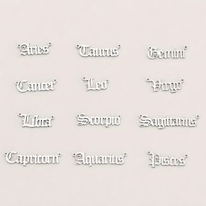 May include: A collection of twelve silver-toned metal charms, each featuring the name of a zodiac sign in an old English gothic font. The charms include Aries, Taurus, Gemini, Cancer, Leo, Virgo, Libra, Scorpio, Sagittarius, Capricorn, Aquarius, and Pisces.
