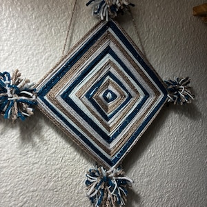 Könnte beinhalten: Ein rautenförmiger Wandbehang aus Garn, das in konzentrischen Quadraten in Marineblau, Weiß und Metallic-Gold gewickelt ist. Das Stück hat vier flauschige Pompons an jeder Ecke und ist an einer dünnen goldenen Schnur aufgehängt.