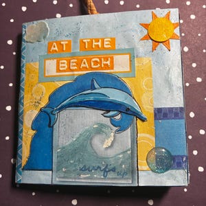 May include: A handmade square art piece with a beach theme. It features a blue dolphin leaping over a wave, with the words "surf's up." The text "AT THE BEACH" is displayed in orange letters. A small orange sun and a ribbon are attached.