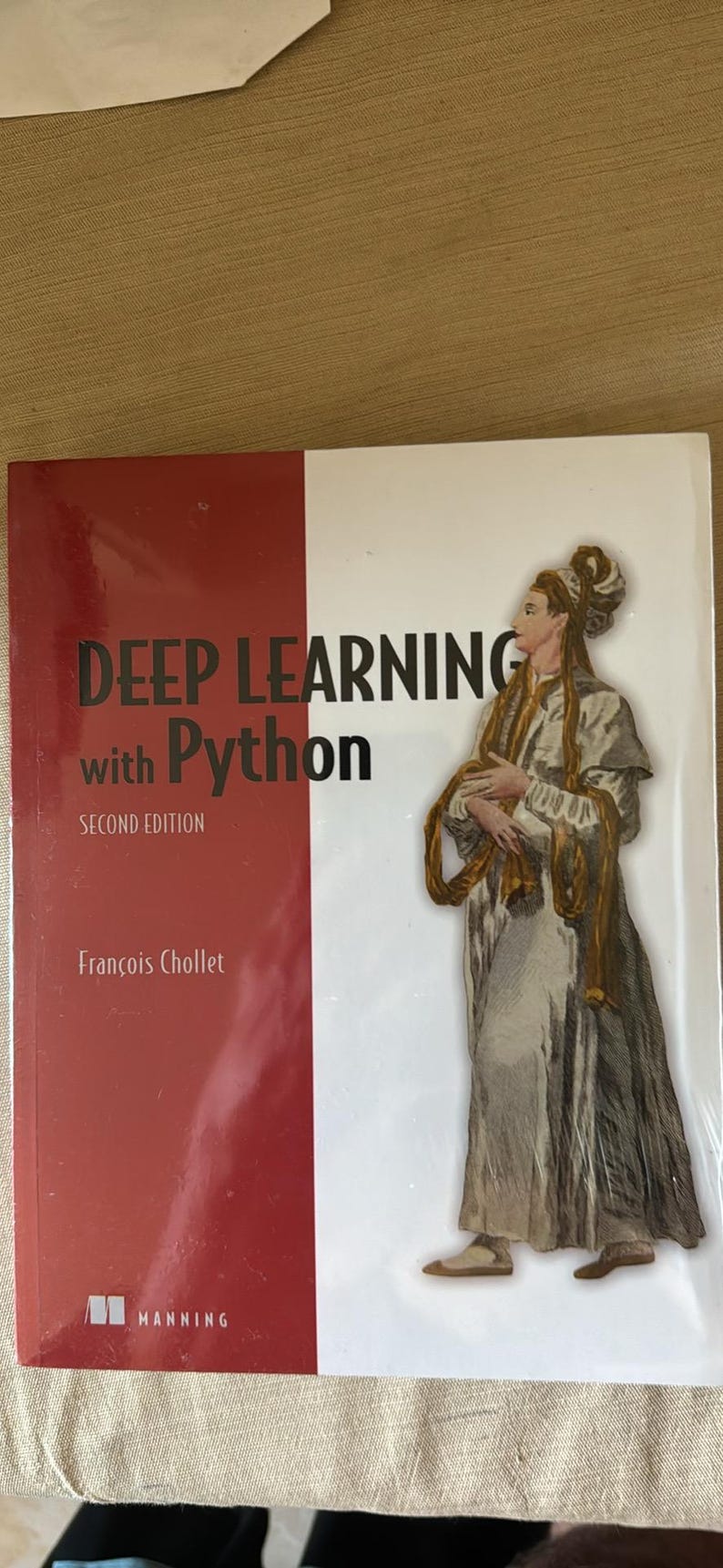May include: The book titled Deep Learning with Python Second Edition by Fran&ccedil;ois Chollet features a red and white cover. The cover displays an illustration of a person in historical attire with long flowing fabric, published by Manning.