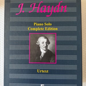 以下が含まれることがあります： 濃い青色の本の表紙。ピンク色の筆記体で「J. Haydn」と書かれ、五線譜の上に配置されています。「Piano Solo Complete Edition」は黄色で表示されています。ピンク色のフレームの中に男性の肖像画があります。「Urtext」の文字は黄色です。