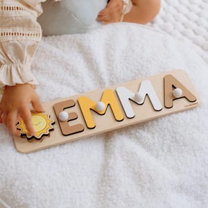 Peut inclure: Puzzle en bois avec le nom "EMMA" en différentes couleurs, dont le marron, le jaune et le blanc. Le puzzle comprend une pièce en forme de soleil et de petits chevilles blanches pour une manipulation facile. Conçu pour les jeunes enfants.