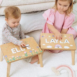 May include: Two small wooden tables with four legs, featuring personalised name puzzles. One table displays the name ELLE with floral graphics, while the other shows the name LAYLA with stars and a rainbow graphic. The tabletops are light wood.