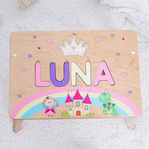May include: A rectangular wooden step stool featuring the name LUNA in large, colorful letters. Above the name is a white crown graphic. Below the name is a rainbow, a cartoon fairy, a castle with pink turrets, and a green dragon holding a flower.