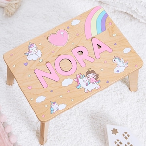 Könnte beinhalten: Ein hellholzfarbener Hocker mit vier Beinen, der den Namen NORA in großen rosa Buchstaben trägt. Die Oberfläche ist mit bunten Illustrationen von Einhörnern, einem Regenbogen, einem rosa Herz und kleinen Wolken auf der Holzmaserung verziert.