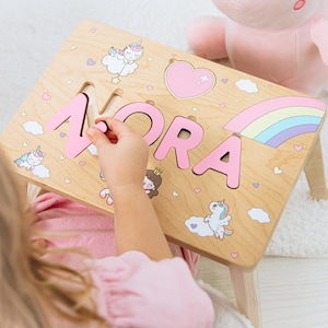 May include: Wooden puzzle table with the name "NORA" in pink letters. The table features illustrations of unicorns, rainbows, and hearts. A pink stuffed dinosaur is in the background.