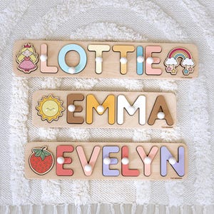 May include: Three wooden name puzzles with peg letters. The top puzzle spells "LOTTIE" with a princess graphic and rainbow. The middle puzzle spells "EMMA" with a sun graphic. The bottom puzzle spells "EVELYN" with a strawberry graphic.