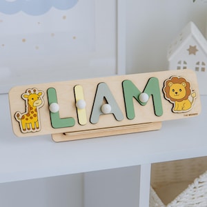 Pu&ograve; includere: Puzzle in legno con il nome "LIAM" con lettere verdi, grigie e gialle. Presenta illustrazioni di una giraffa e di un leone. Il puzzle &egrave; su una mensola bianca. Il testo "LITTLE STAR" &egrave; visibile sullo sfondo.