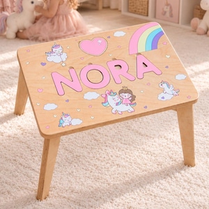 Könnte beinhalten: Ein heller Holztisch für den Schoß mit dem Namen NORA in großen rosa Buchstaben. Die Oberfläche ist mit bunten Illustrationen von Einhörnern, einem Regenbogen, einem rosa Herz und kleinen Wolken verziert und steht auf vier stabilen Holzbeinen.