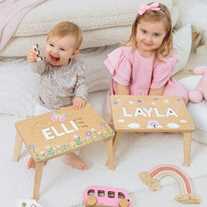 May include: Two light-colored wooden activity tables featuring personalized names in white block letters. One table displays the name ELLIE with a butterfly and floral design, while the other shows the name LAYLA with stars, clouds, and a rainbow graphic.