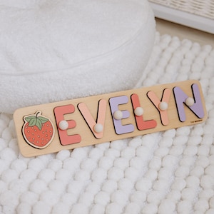 Pu&ograve; includere: Puzzle con nome in legno con la scritta "EVELYN" in lettere dai colori pastello e pomelli bianchi. Una grafica a fragola &egrave; a sinistra. Il puzzle &egrave; su una superficie bianca strutturata.