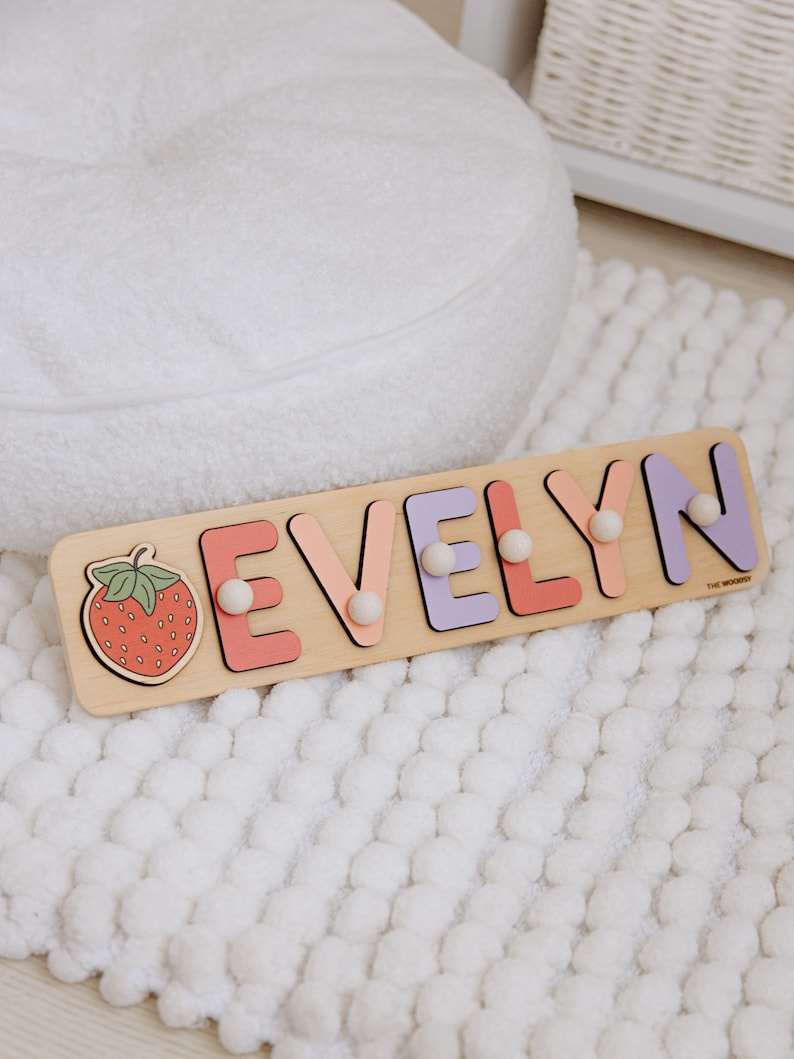 Pu&ograve; includere: Puzzle con nome in legno con la scritta "EVELYN" in lettere dai colori pastello e pomelli bianchi. Una grafica a fragola &egrave; a sinistra. Il puzzle &egrave; su una superficie bianca strutturata.