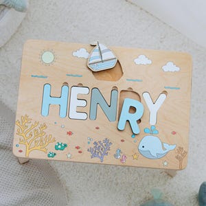 Puede incluir: Un taburete de madera con un rompecabezas de nombre personalizado que presenta el nombre "HENRY" en letras azules, blancas y grises. El taburete tiene un tema náutico con un velero, una ballena y un arrecife de coral.