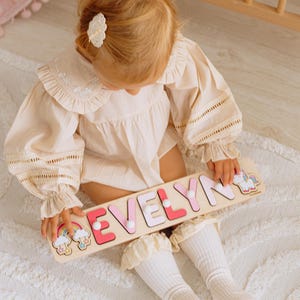 Pu&ograve; includere: Un puzzle di legno con il nome "EVELYN" in lettere rosa e bianche, con illustrazioni di arcobaleni e unicorni. Il puzzle &egrave; tenuto in mano da un bambino che indossa un completo color crema e calzini bianchi.