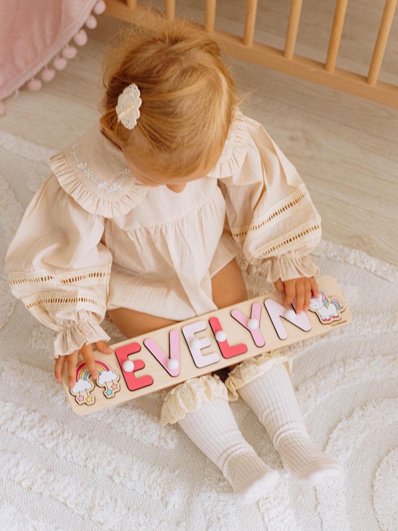 Pu&ograve; includere: Un puzzle di legno con il nome "EVELYN" in lettere rosa e bianche, con illustrazioni di arcobaleni e unicorni. Il puzzle &egrave; tenuto in mano da un bambino che indossa un completo color crema e calzini bianchi.