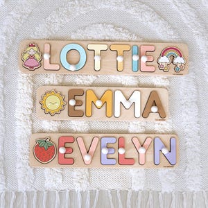 Pu&ograve; includere: Tre puzzle di nomi in legno con i nomi "Lottie", "Emma" ed "Evelyn". Ogni puzzle presenta lettere colorate ed elementi decorativi come una principessa, un sole, un arcobaleno e una fragola. I puzzle sono su una superficie bianca strutturata.
