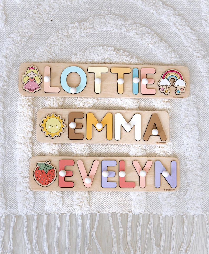Pu&ograve; includere: Tre puzzle di nomi in legno con i nomi "Lottie", "Emma" ed "Evelyn". Ogni puzzle presenta lettere colorate ed elementi decorativi come una principessa, un sole, un arcobaleno e una fragola. I puzzle sono su una superficie bianca strutturata.