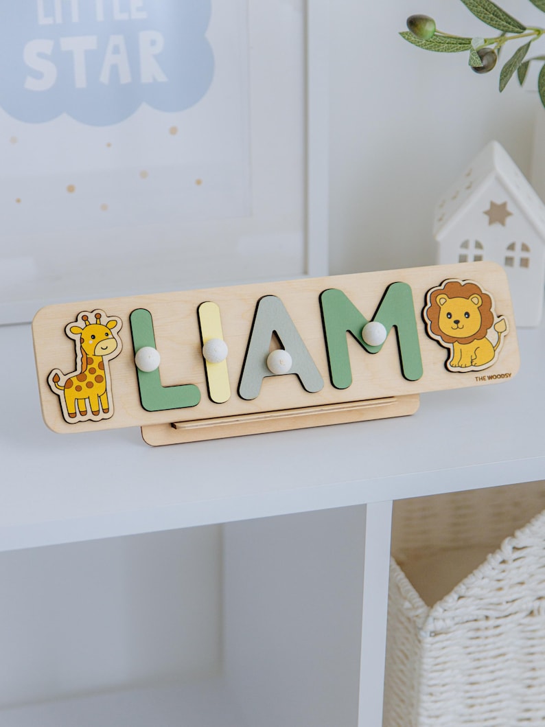 Pu&ograve; includere: Puzzle in legno con il nome "LIAM" con lettere verdi, grigie e gialle. Presenta illustrazioni di una giraffa e di un leone. Il puzzle &egrave; su una mensola bianca. Il testo "LITTLE STAR" &egrave; visibile sullo sfondo.