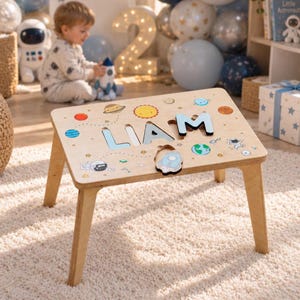 May include: A light-colored wooden activity table featuring a space-themed design with colorful planets, stars, and astronauts. The tabletop includes a cutout puzzle area with the name LIAM spelled out in large, removable wooden letters.