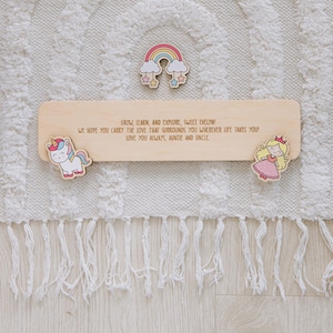 Pu&ograve; includere: Cartello in legno con la scritta "GROW, LEARN, AND EXPLORE, SWEET EVELYN!" e un arcobaleno, un unicorno e un design a principessa. Il cartello &egrave; circondato da un tappeto bianco con frange.